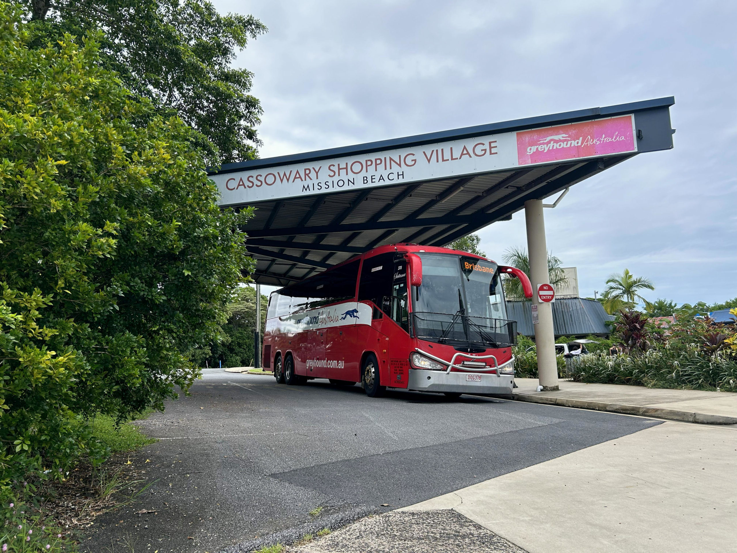 Greyhound Bus at the Bus Stop in Wongaling Beach. You can see the Bus it self, parked under the cover of the Cassowary Shopping Village in Mission Beach. The Sky is cloudy grey. In the background, you can see the shopping village.